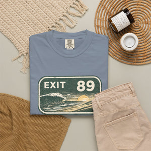 A folded light blue t-shirt with a retro 'EXIT 13' beach sunset graphic, styled in a flat lay with beige pants, a woven blanket, a candle jar, and a small cup on a neutral background.