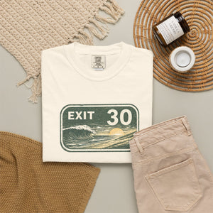 A folded cream T-shirt with a vintage-style 'EXIT 30' road sign graphic featuring a wave and sun, styled in a flat lay with beige pants, a woven mat, a candle jar, and a small cup on a light gray surface.