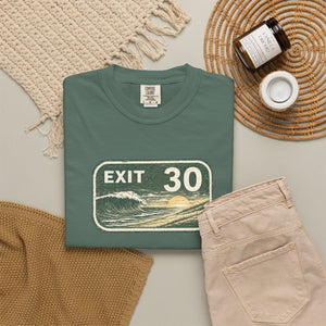 A folded sage green graphic T-shirt is arranged in a styled flat lay on a light gray surface, with a beige woven scarf, tan knit fabric, folded beige pants, a small amber candle jar, and a white cup nearby. The shirt has a large front graphic reading “EXIT 30” over a wave and sun illustration.