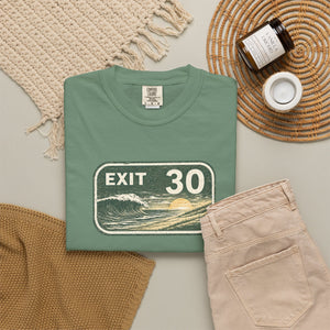 A folded sage green t-shirt with a retro 'EXIT 30' wave graphic, styled in a flat lay with beige pants, a woven blanket, a candle jar, and a small cup on a neutral background.
