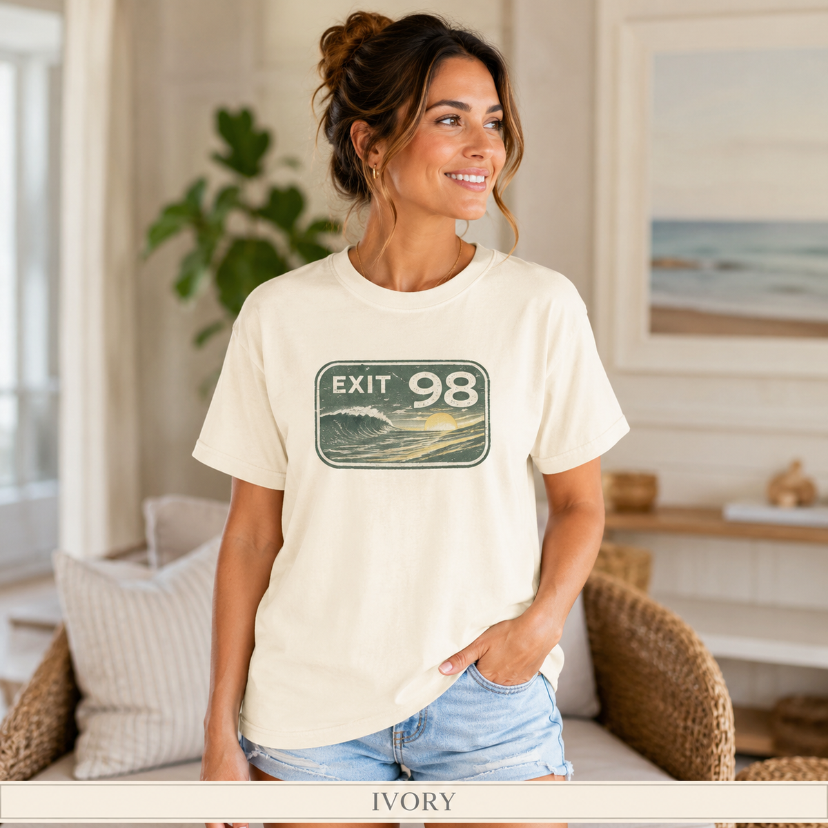 A woman wearing an ivory short-sleeve graphic t-shirt with a retro 'EXIT 98' wave design, paired with light-wash denim shorts in a bright, modern indoor setting.