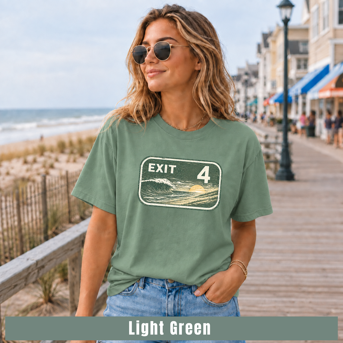 A woman wearing a light green graphic t-shirt with a retro 'EXIT 4' wave sign design, standing on a wooden boardwalk next to the ocean. A text banner at the bottom reads 'Light Green'.