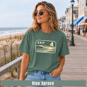 A woman wearing a muted blue-green graphic T-shirt with a retro 'EXIT 4' sign design, standing on a boardwalk near the beach. The bottom of the image has a color label reading 'Blue Spruce'.