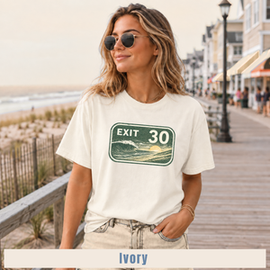 A woman wearing round sunglasses and a cream-colored graphic t-shirt with an 'EXIT 30' ocean sign design, standing on a wooden boardwalk next to the ocean. A label at the bottom reads 'Ivory'.