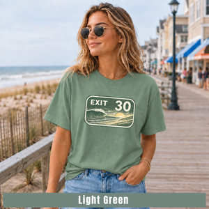 A woman wearing a light green graphic t-shirt with a 'EXIT 30' sunset sign design, standing on a wooden boardwalk next to the ocean. A text banner at the bottom reads 'Light Green'.