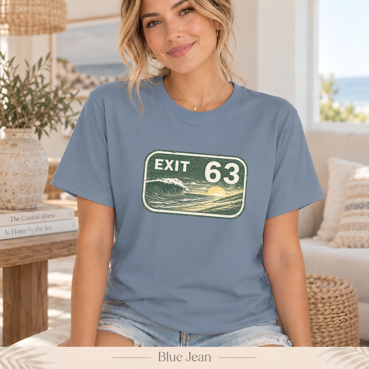 A woman wearing a blue graphic t-shirt with a 'EXIT 63' ocean wave design, seated in a bright, modern living room setting.