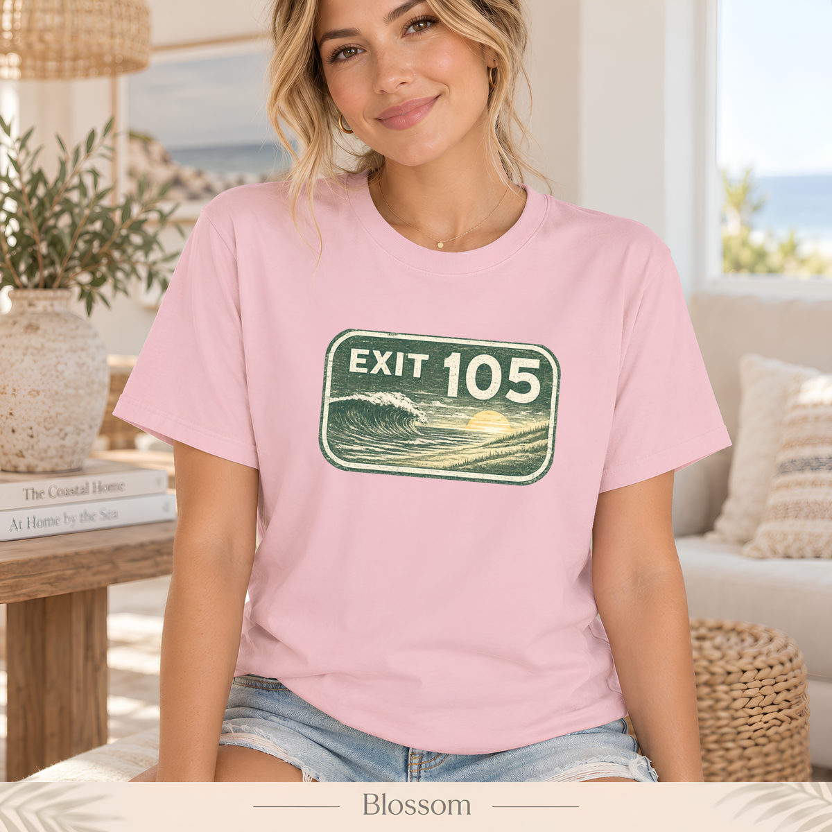 A woman wearing a light pink crewneck t-shirt with a green 'EXIT 105' wave graphic on the chest, sitting in a bright, modern living room. The bottom of the image features a banner with the text 'Blossom'.