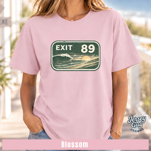 A woman wearing a light pink t-shirt with a retro 'EXIT 89' highway sign graphic on the chest. The shirt is labeled as 'Blossom' by Jersey Guy Designs, shown in an outdoor setting with a blurred background.