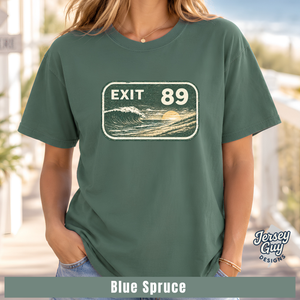 A woman wearing a muted blue-green graphic T-shirt with a retro 'EXIT 89' ocean wave print on the chest. The image includes a 'Jersey Guy Designs' logo and a text banner at the bottom reading 'Blue Spruce'.