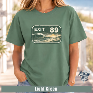 A woman wearing a light green graphic t-shirt with a retro 'EXIT 89' ocean wave design, shown in an outdoor setting with a 'Jersey Guy Designs' logo overlay.