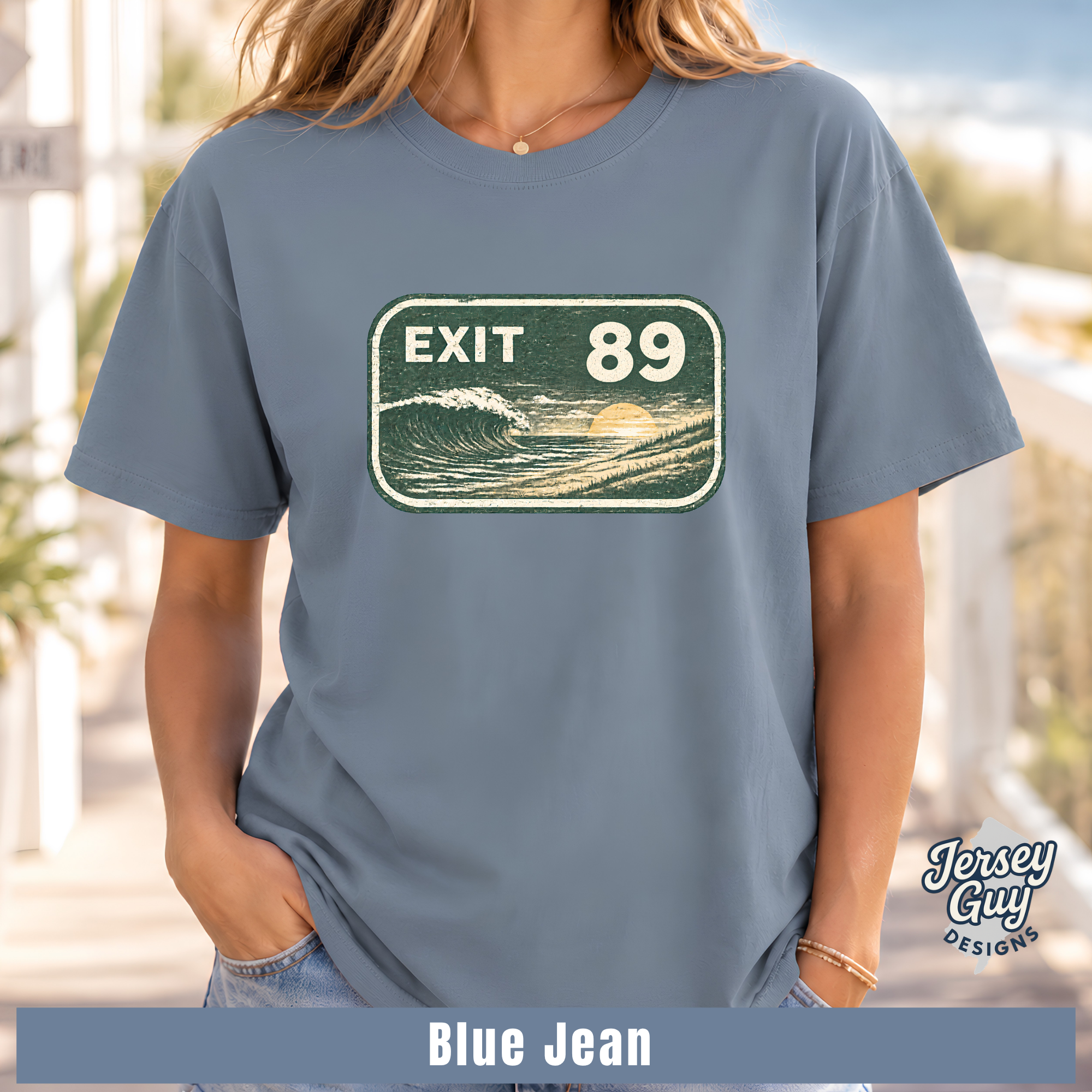 A woman wearing a slate blue graphic t-shirt with a retro 'EXIT 89' wave sign design. The image has a 'Blue Jean' color label at the bottom and a 'Jersey Guy Designs' logo on the right.