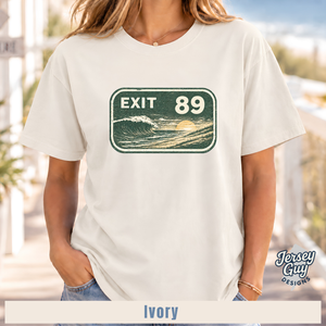 A woman wearing an ivory short-sleeve t-shirt with a distressed green and white 'EXIT 89' ocean wave graphic on the chest. The background is a blurred outdoor beach setting, and a 'Jersey Guy Designs' brand logo is visible in the bottom right corner.