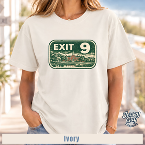 A woman wearing an ivory short-sleeve t-shirt with a distressed green and white 'EXIT 9' sign graphic on the front. The background is a softly blurred outdoor setting.