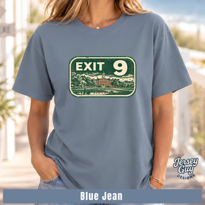 A woman wearing a slate blue graphic t-shirt with a distressed green and white 'EXIT 9' sign design on the front. The image has a 'Blue Jean' color label at the bottom and a 'Jersey Guy Designs' logo on the right.