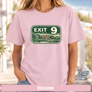 A woman wearing a light pink short-sleeve t-shirt with a vintage-style green 'EXIT 9' sign graphic on the chest. The image is a lifestyle mockup with a blurred outdoor background, featuring a 'Jersey Guy Designs' logo and a pink banner at the bottom labeled 'Blossom'.