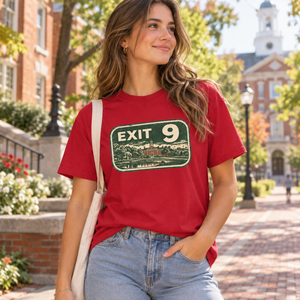 A woman wearing a red graphic t-shirt with a vintage-style green 'EXIT 9' sign print, paired with light-wash blue jeans and a cream shoulder bag, standing on a sunny brick campus walkway.