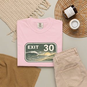 A folded light pink T-shirt with a retro 'EXIT 30' road sign graphic featuring a wave and sunset, styled in a flat lay with beige pants, a woven blanket, a candle jar, and a small cup on a neutral background.