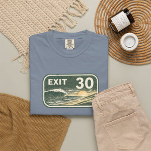 A folded dusty blue t-shirt with a retro 'EXIT 30' ocean wave graphic, styled in a flat lay with beige pants, a woven blanket, a candle, and a small cup on a neutral background.