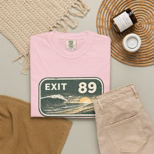 A folded light pink T-shirt with a retro 'EXIT 89' road sign graphic, styled in a flat lay with beige pants, a woven blanket, a candle jar, and a small cup on a neutral background.