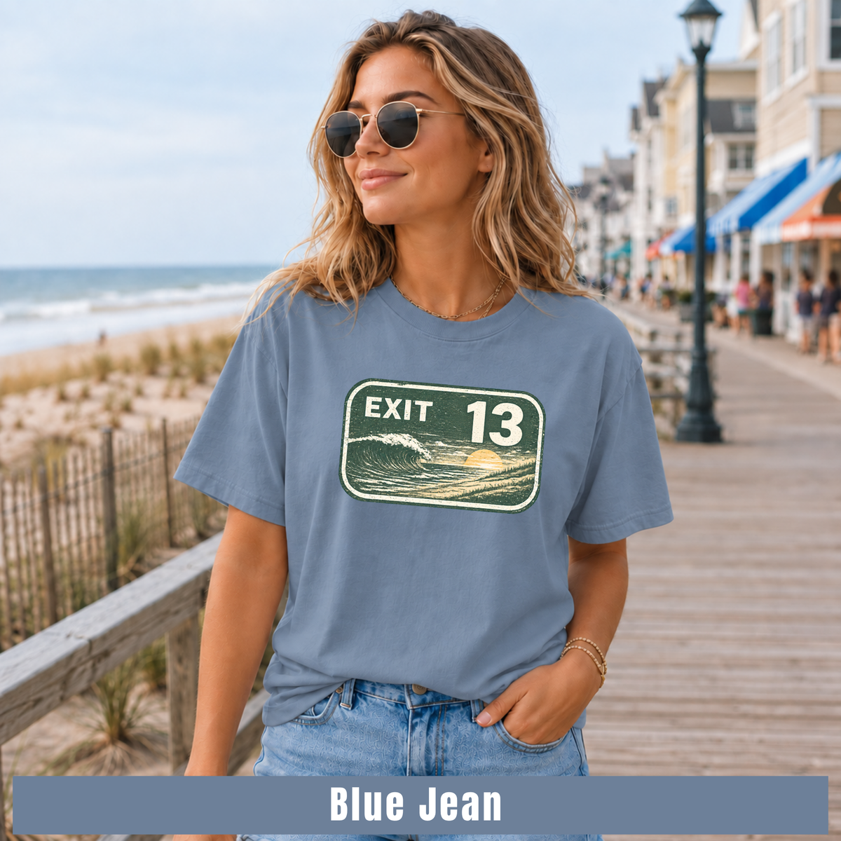 A woman wearing a blue graphic t-shirt with an 'EXIT 13' ocean sunset print, standing on a wooden boardwalk next to the ocean. A text banner at the bottom reads 'Blue Jean'.