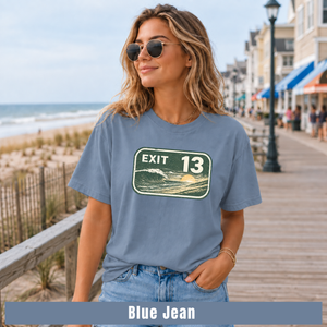A woman wearing a blue graphic t-shirt with an 'EXIT 13' ocean sunset print, standing on a wooden boardwalk next to the ocean. A text banner at the bottom reads 'Blue Jean'.