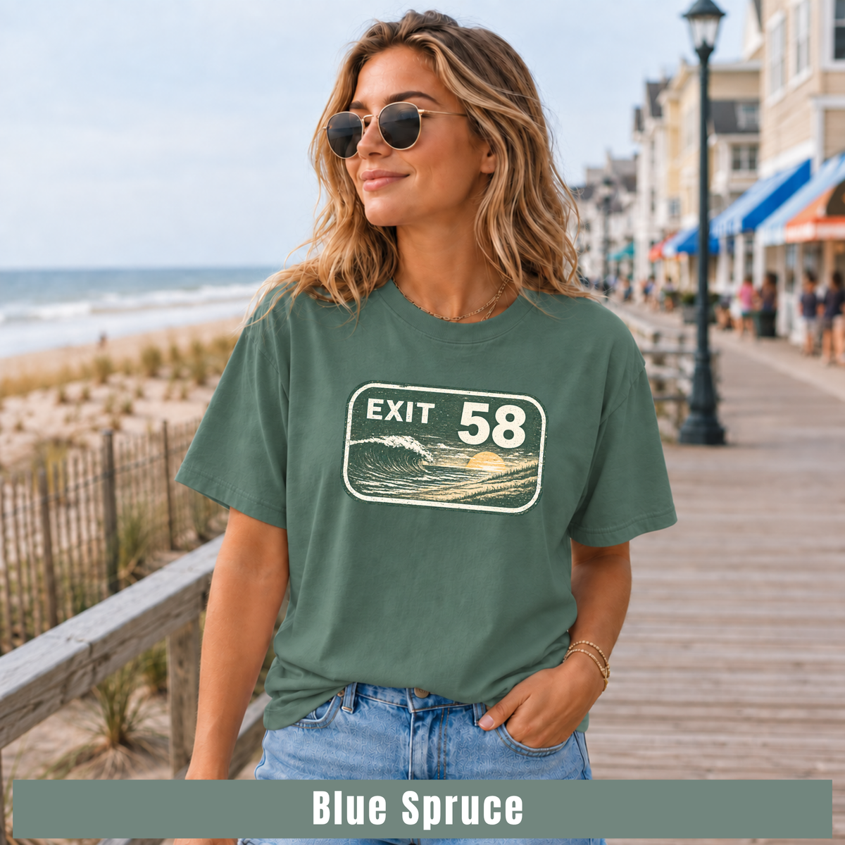 A woman wearing a muted blue-green graphic t-shirt with a retro 'EXIT 58' wave design, standing on a wooden boardwalk next to the ocean. The bottom of the image features a color label overlay that reads 'Blue Spruce'.